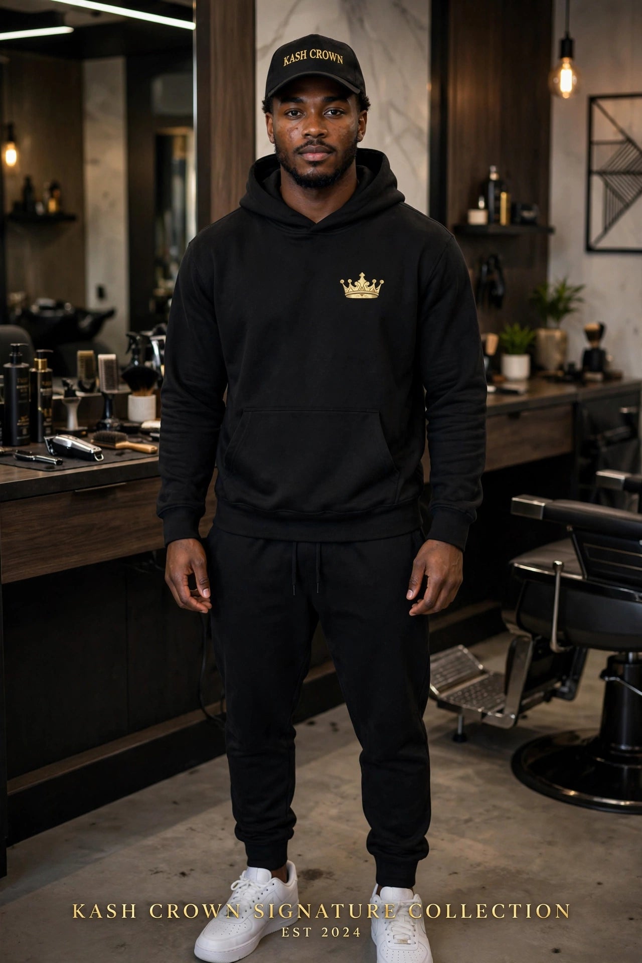 A man wearing a black hoodie with a gold crown logo on the chest, black joggers, and a black cap with 'KASH CROWN' text, standing in a modern barbershop environment.
