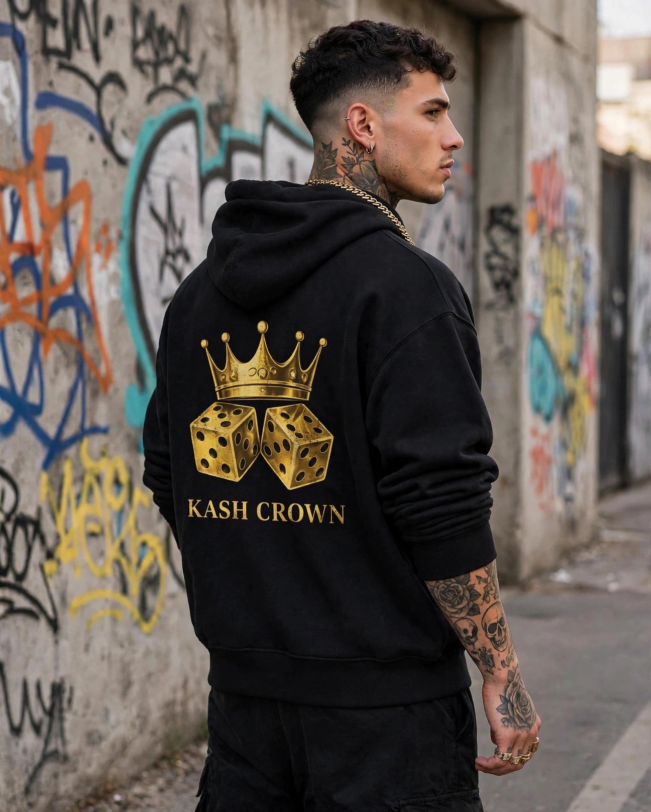 Back view of the black KASH CROWN Crown & Dice Hoodie worn beside a graffiti wall, gold crown and dice graphic above the KASH CROWN wordmark