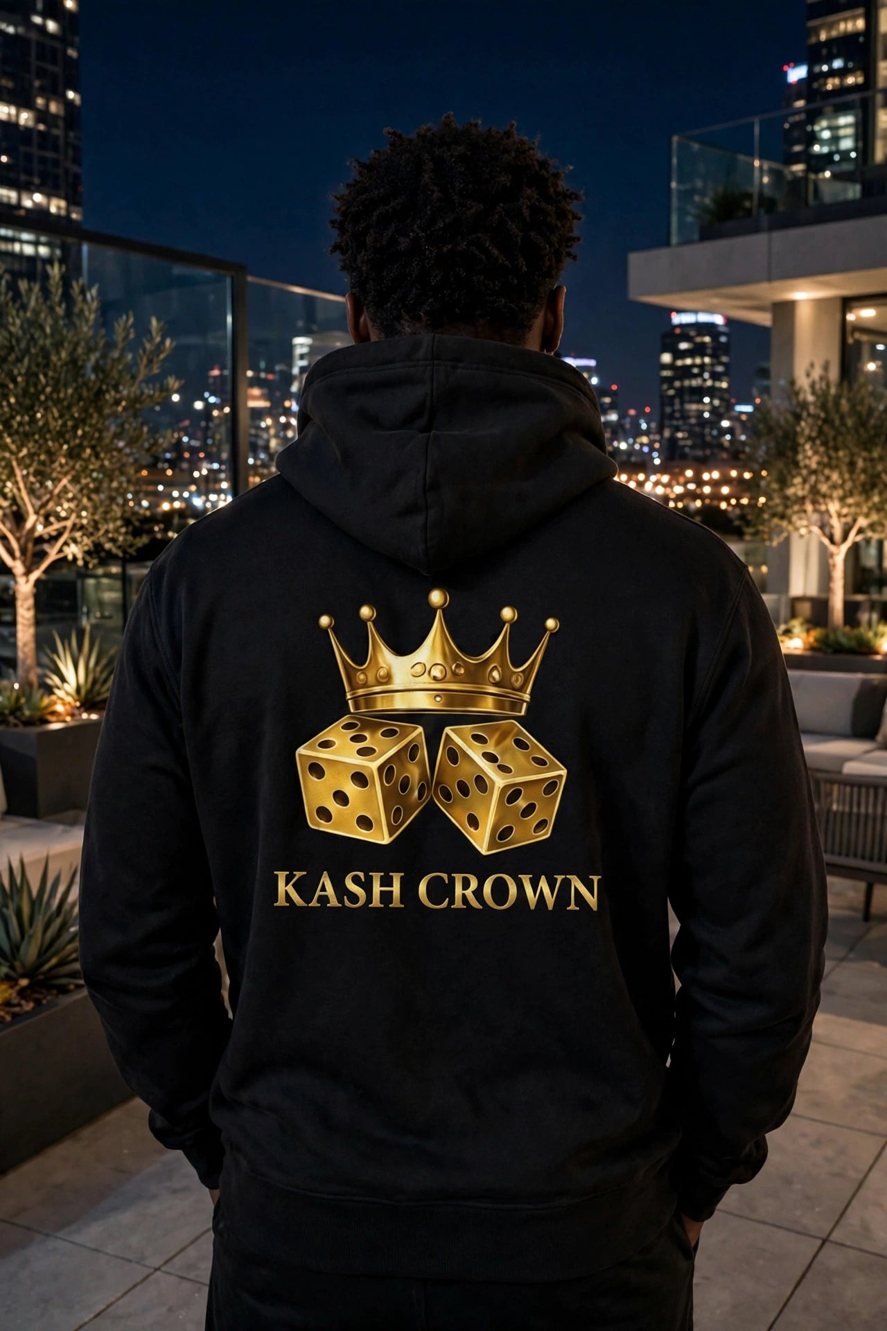 Back view of the black KASH CROWN Crown & Dice Hoodie on a rooftop at night with a city skyline behind, gold crown and dice graphic